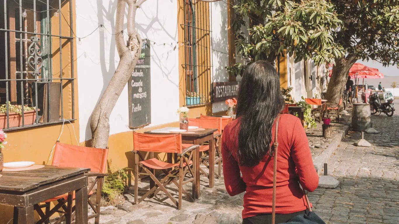 Mulher com blusa vermelha caminhando por uma rua de Colonia del Sacramento com várias cadeiras e mesas na calçada.