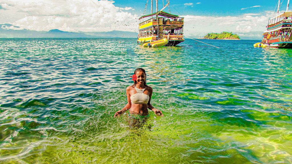 Mulher com uma flor vermelha nos cabelos jogando água para cima com as mãos em uma praia de Paraty, de águas verdes, ao fundo uma escuna se funde no horizonte com o céu azul.