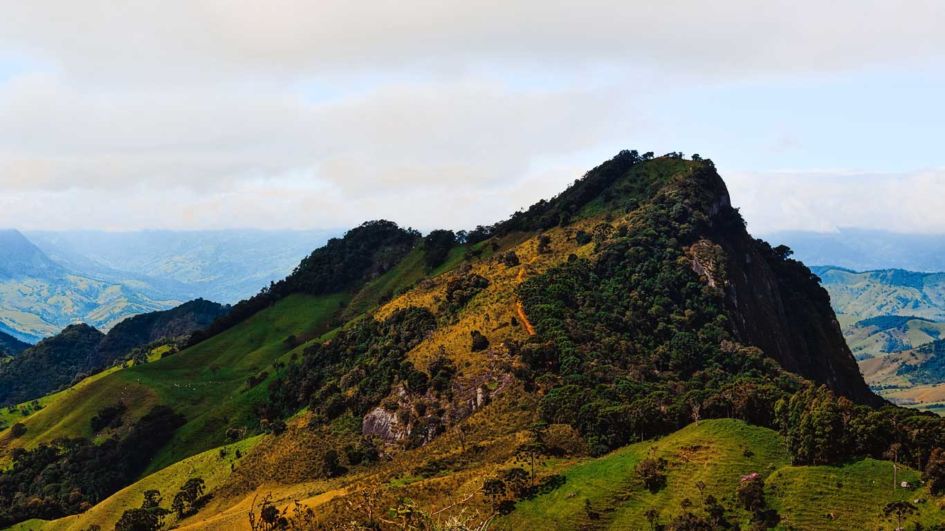 Paisagem montanhosa de Gonçalves com colinas verdejantes e uma formação rochosa proeminente ao fundo. O céu parcialmente nublado cobre o cenário, e as montanhas distantes são visíveis no horizonte.