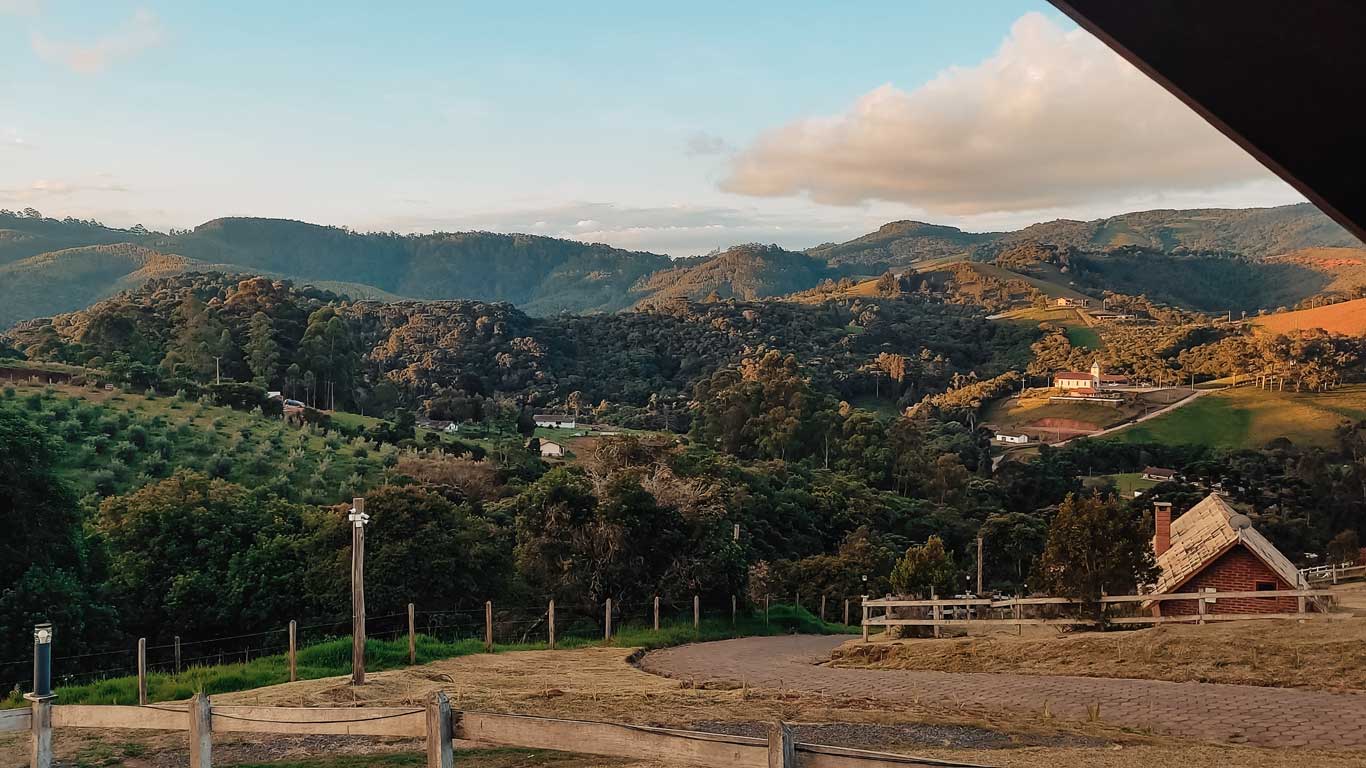 Vista panorâmica das montanhas e vales de Monte Verde, uma das cidades próximas a Campos do Jordão, com vegetação densa, colinas suaves e pequenas casas ao longe, sob um céu parcialmente nublado ao pôr do sol.