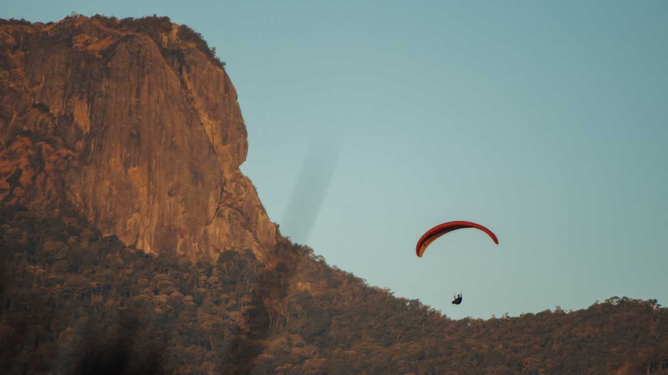 Uma paisagem montanhosa em São Bento do Sapucaí com um praticante de parapente no ar, voando perto de um grande paredão de pedra, sob um céu azul claro.