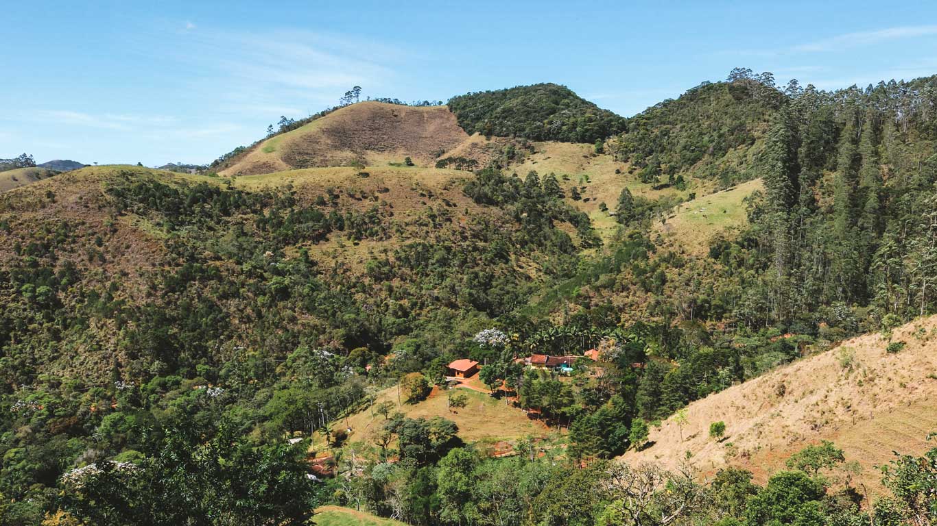 Paisagem rural de São Francisco Xavier, com uma mistura de árvores altas e colinas verdes, e pequenas casas espalhadas pela região montanhosa sob um céu azul