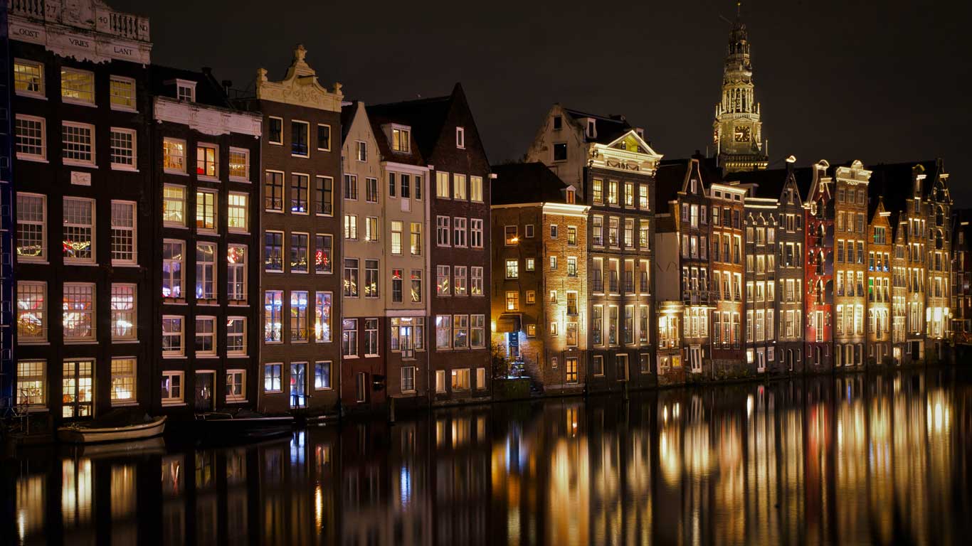 Casas tradicionais de frente para o canal em Amsterdã, iluminadas à noite com reflexos coloridos na água. A arquitetura característica e a iluminação suave destacam a beleza histórica da cidade. Ao fundo, uma torre de igreja se eleva contra o céu noturno.