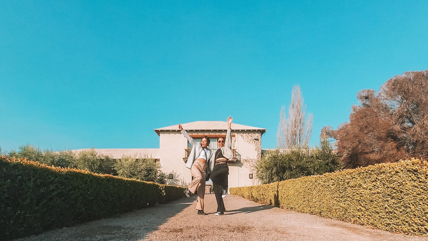 Duas mulheres posando alegremente em frente a uma vinícola em Mendoza, Argentina, com arbustos bem cuidados ao longo do caminho que leva à entrada do edifício. O céu está claro e azul, complementando o cenário tranquilo e ensolarado.