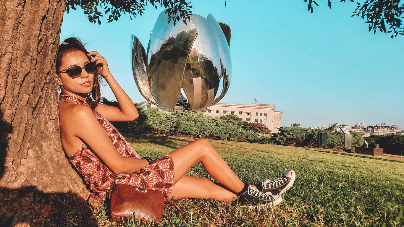 Mulher sentada à sombra de uma árvore, usando óculos escuros e vestido vermelho, com a famosa escultura Floralis Genérica ao fundo em Buenos Aires. O dia está ensolarado e a paisagem verde do parque contrasta com o céu azul claro.