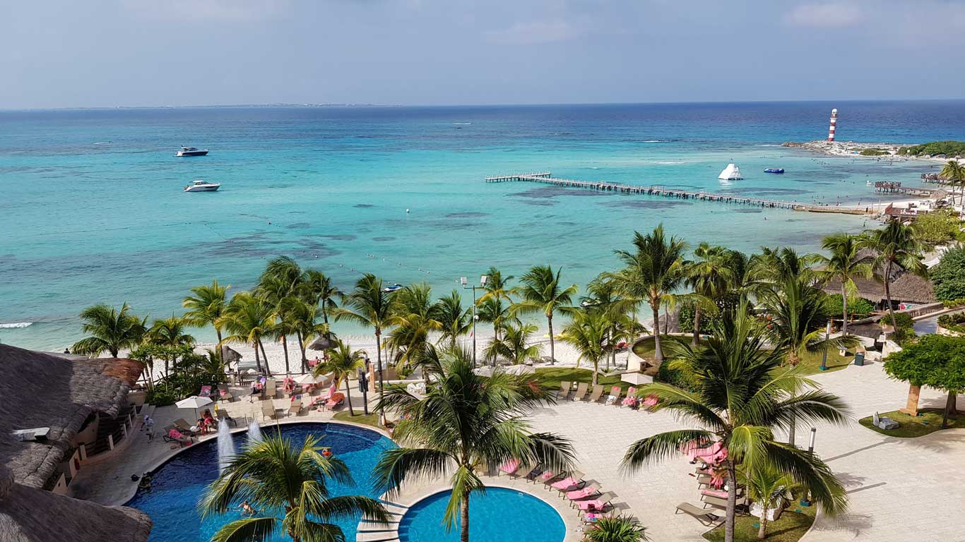 Vista aérea de uma praia paradisíaca em Cancún, México, com águas cristalinas azul-turquesa, várias palmeiras, espreguiçadeiras ao redor de uma piscina e barcos ancorados no mar. Ao fundo, há um píer que se estende até o oceano e um farol na ponta da península, caracterizando um dos melhores lugares para viajar com amigos.