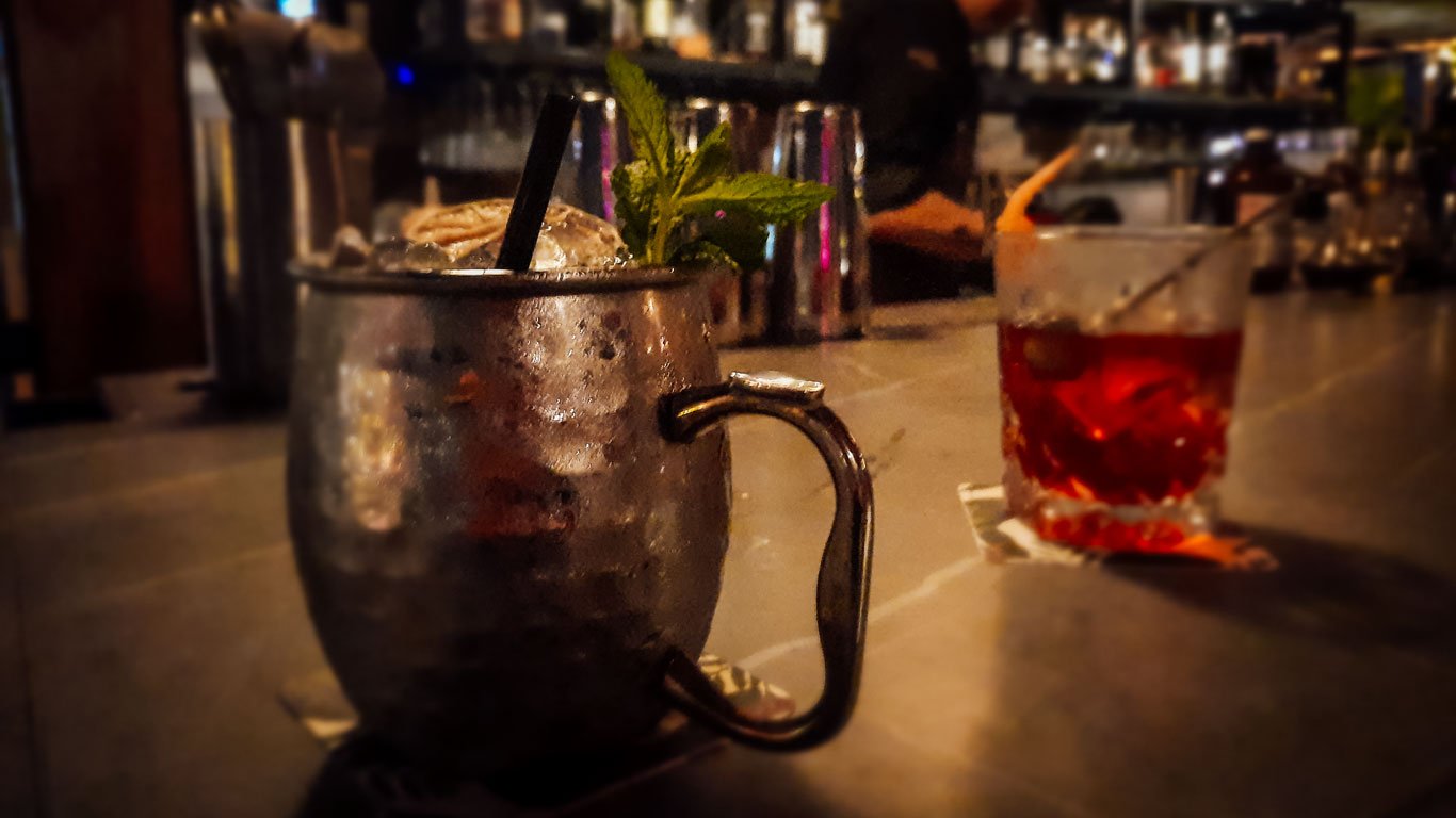 Dois coquetéis em um bar em Cartagena, com destaque para uma caneca de Moscow Mule adornada com folhas de hortelã e um copo baixo com uma bebida vermelha. O ambiente ao fundo é acolhedor e intimista, perfeito para desfrutar de uma noite na cidade.