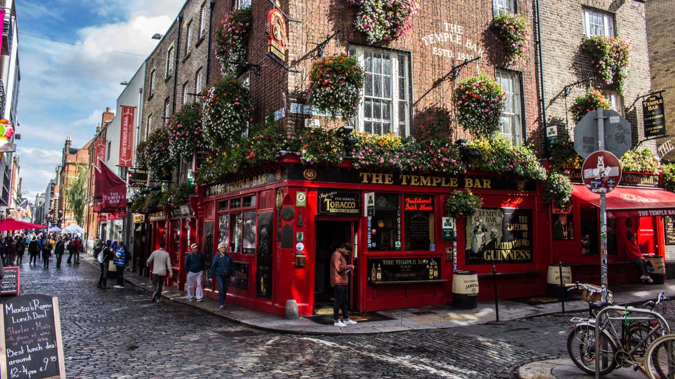 Fachada do famoso pub The Temple Bar em Dublin, com sua vibrante pintura vermelha e cestas de flores coloridas penduradas nas janelas. A rua de paralelepípedos ao redor está movimentada com pedestres explorando a área histórica e cultural da cidade.