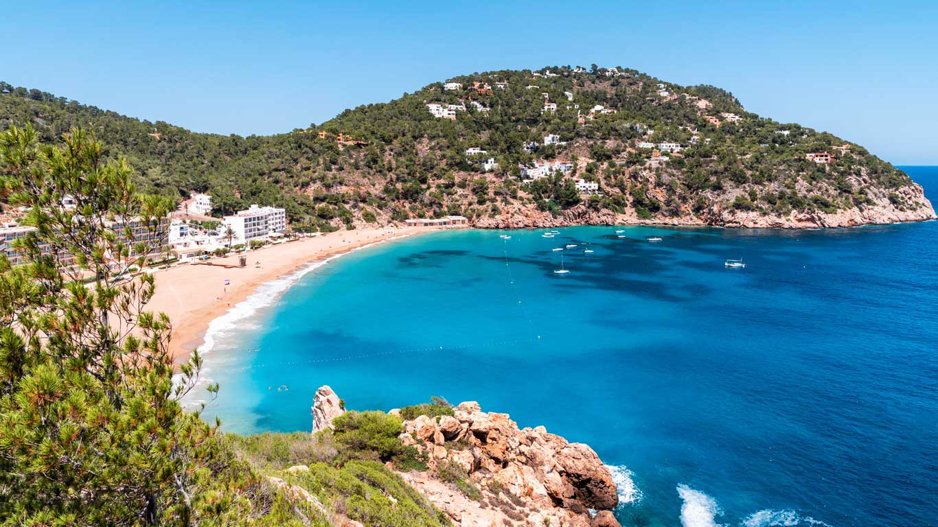 Vista panorâmica de uma praia de areia dourada em Ibiza, Espanha, cercada por colinas verdes e águas cristalinas azul-turquesa. Vários barcos estão ancorados na baía tranquila, e há edificações brancas ao longo da costa.