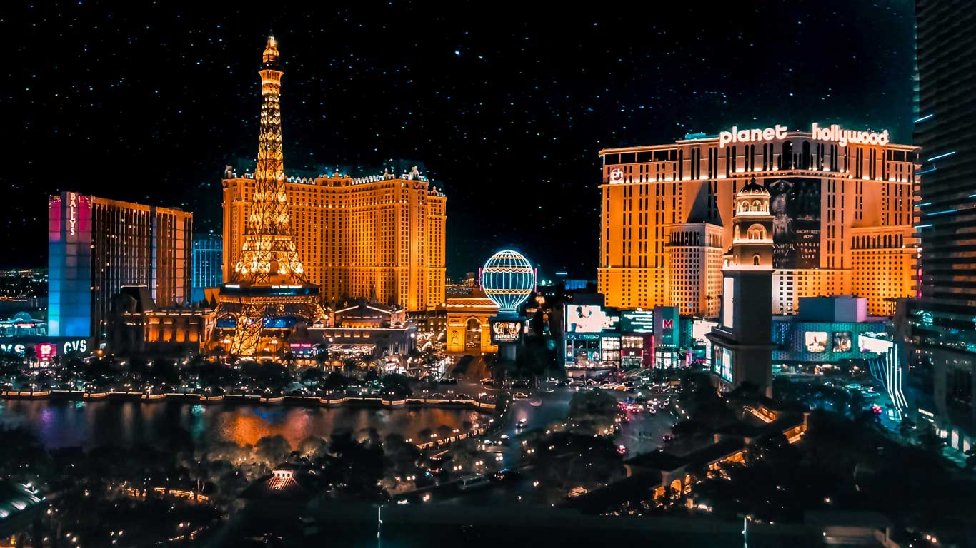 Vista noturna da Las Vegas Strip, com destaque para o hotel Paris Las Vegas e sua réplica iluminada da Torre Eiffel. O hotel Planet Hollywood e outros cassinos icônicos brilham ao fundo, sob um céu estrelado, mostrando a vibrante vida noturna da cidade.