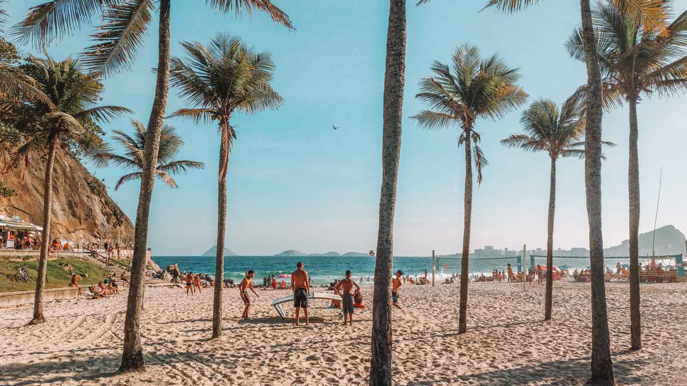 Praia do Leme no Rio de Janeiro, com pessoas aproveitando o dia ensolarado entre palmeiras e areia clara. Grupos jogam vôlei e frescobol, enquanto outros relaxam à sombra. O mar azul e o céu claro complementam o cenário vibrante e animado.