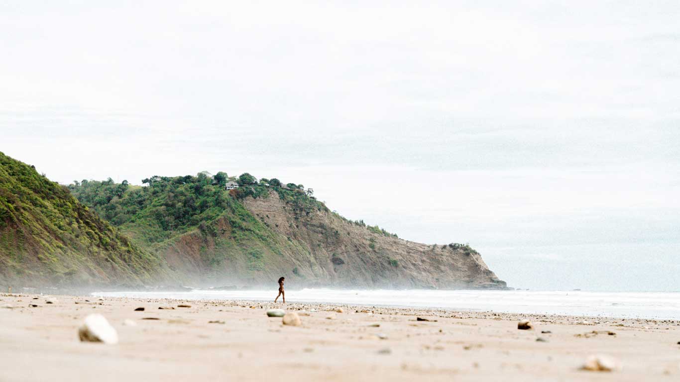 Paisagem tranquila da praia de Montañita, Equador, com uma pessoa caminhando sozinha na areia, cercada por falésias verdes. O céu está nublado, criando uma atmosfera serena e isolada.