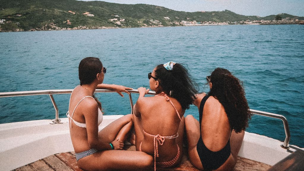 Três mulheres de biquíni sentadas na borda de um barco, apreciando a vista do mar azul em Arraial do Cabo. Ao fundo, vê-se a costa verdejante com algumas casas espalhadas pelas colinas. O cenário é perfeito para um passeio relaxante e ensolarado.