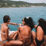 Três mulheres de biquíni sentadas na borda de um barco, apreciando a vista do mar azul em Arraial do Cabo. Ao fundo, vê-se a costa verdejante com algumas casas espalhadas pelas colinas. O cenário é perfeito para um passeio relaxante e ensolarado.