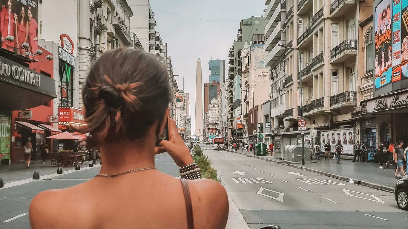 Mulher vista de costas fotografando a movimentada Avenida Corrientes em Buenos Aires, Argentina, com o icônico Obelisco ao fundo, destacando a vibrante vida urbana e pontos arquitetônicos marcantes.