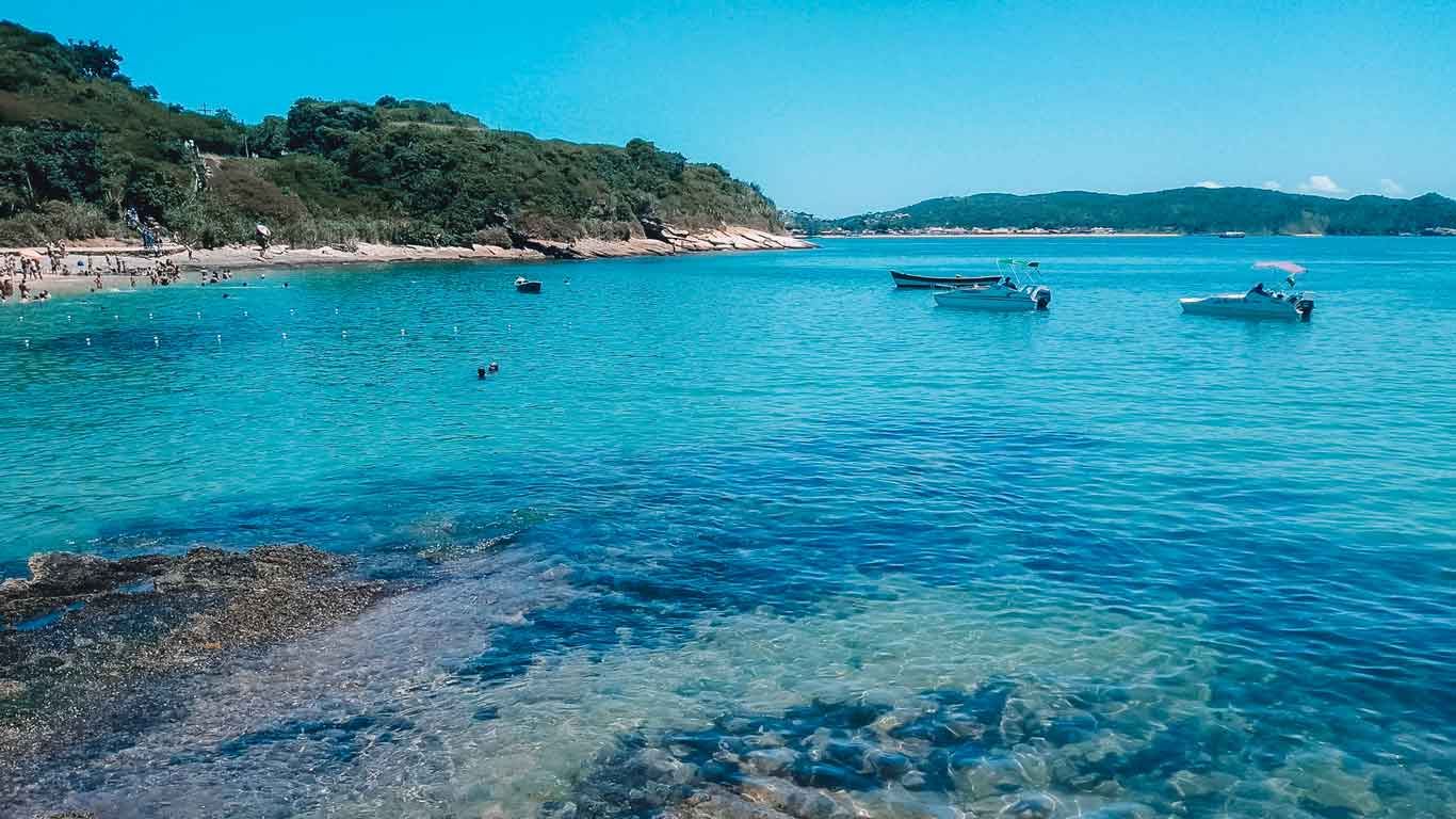 Imagem da Praia de João Fernandes em Búzios, com águas transparentes e alguns barcos ao fundo.