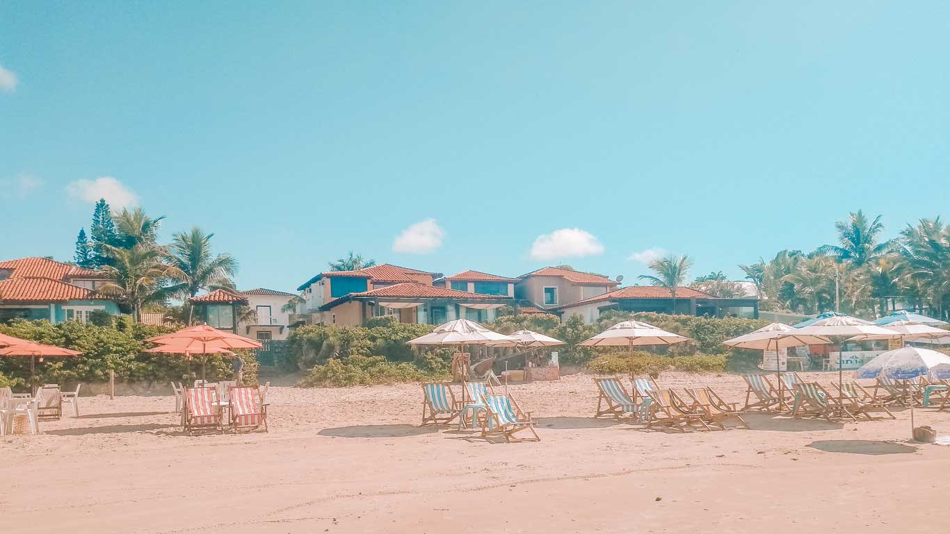 Uma cena da Praia de Geribá, uma excelente opção de onde ficar em Búzios, com diversas cadeiras na areia da praia com casas ao fundo.