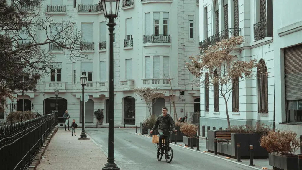 A imagem captura um momento tranquilo em Montevidéu, mostrando um homem pedalando sua bicicleta ao longo de uma rua ladeada por prédios históricos de arquitetura europeia. Árvores de folhas esparsas e luminárias de estilo clássico adicionam charme ao cenário, enquanto uma cerca metálica delimita um dos lados da via.