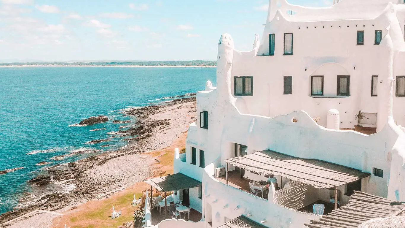 Estrutura arquitetônica branca e única do Hotel Casapueblo, com curvas orgânicas, com vista para um mar azul calmo, acompanhada de uma frente de praia rochosa. Móveis brancos para áreas externas estão dispostos em terraços, sugerindo uma atmosfera serena de resort litorâneo.