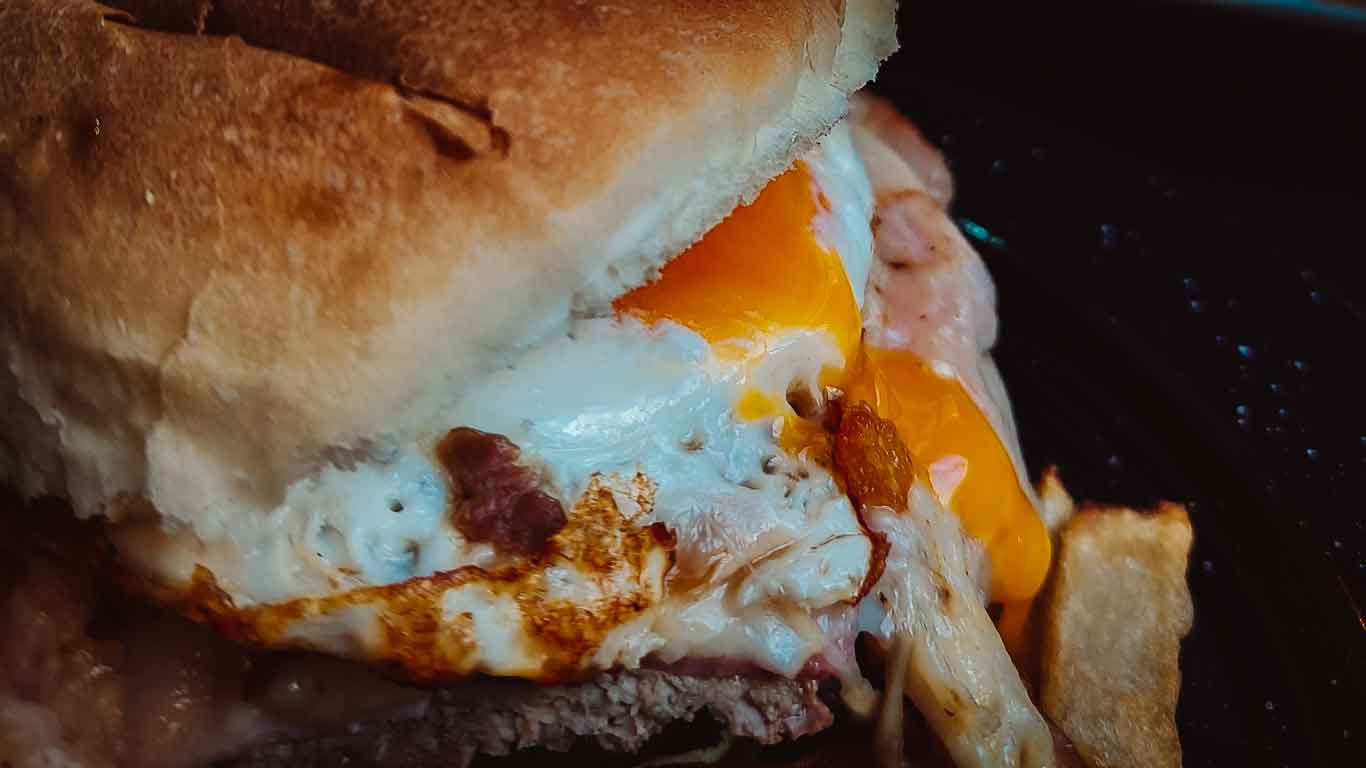 Imagem do típico sanduíche uruguaio, o chivito, onde a gema de ovo escorre delicadamente sobre os demais ingredientes, realçando o apetitoso conjunto de sabores.