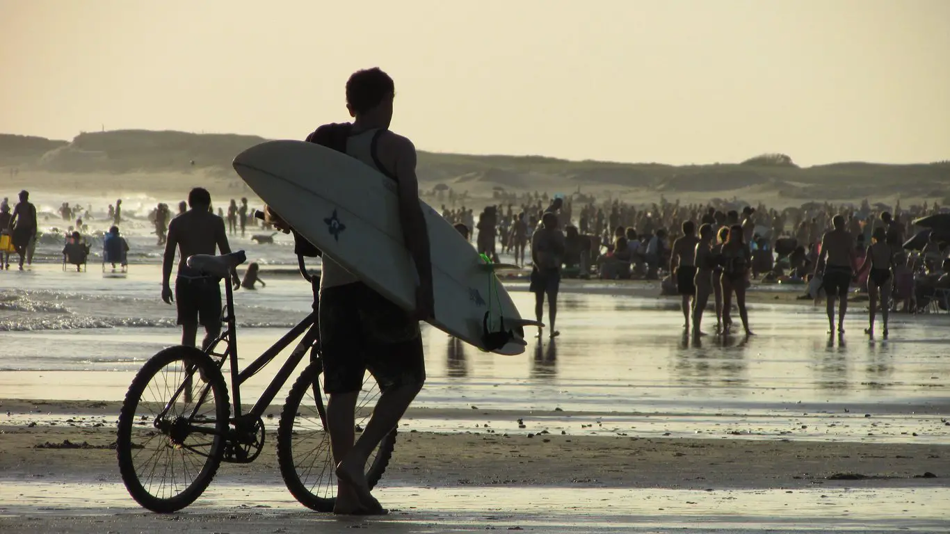 Praia movimentada em La Paloma, um dos melhores lugares para conhecer no Uruguai, ao entardecer, com um grande número de banhistas caminhando e aproveitando a faixa de areia molhada. Em primeiro plano, um homem caminha empurrando uma bicicleta com uma prancha de surfe debaixo do braço, destacando o estilo de vida praiano e esportivo da região.