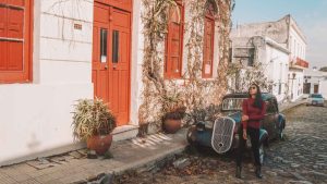 Uma mulher com óculos de sol, vestindo uma blusa vermelha de mangas compridas, está encostada em um carro antigo em uma rua de paralelepípedos. Ao fundo, vê-se uma construção de paredes brancas com portas e janelas vermelhas e plantas trepadeiras secas nas paredes. O cenário é de uma área histórica, possivelmente em uma cidade colonial, com um clima tranquilo e ensolarado.