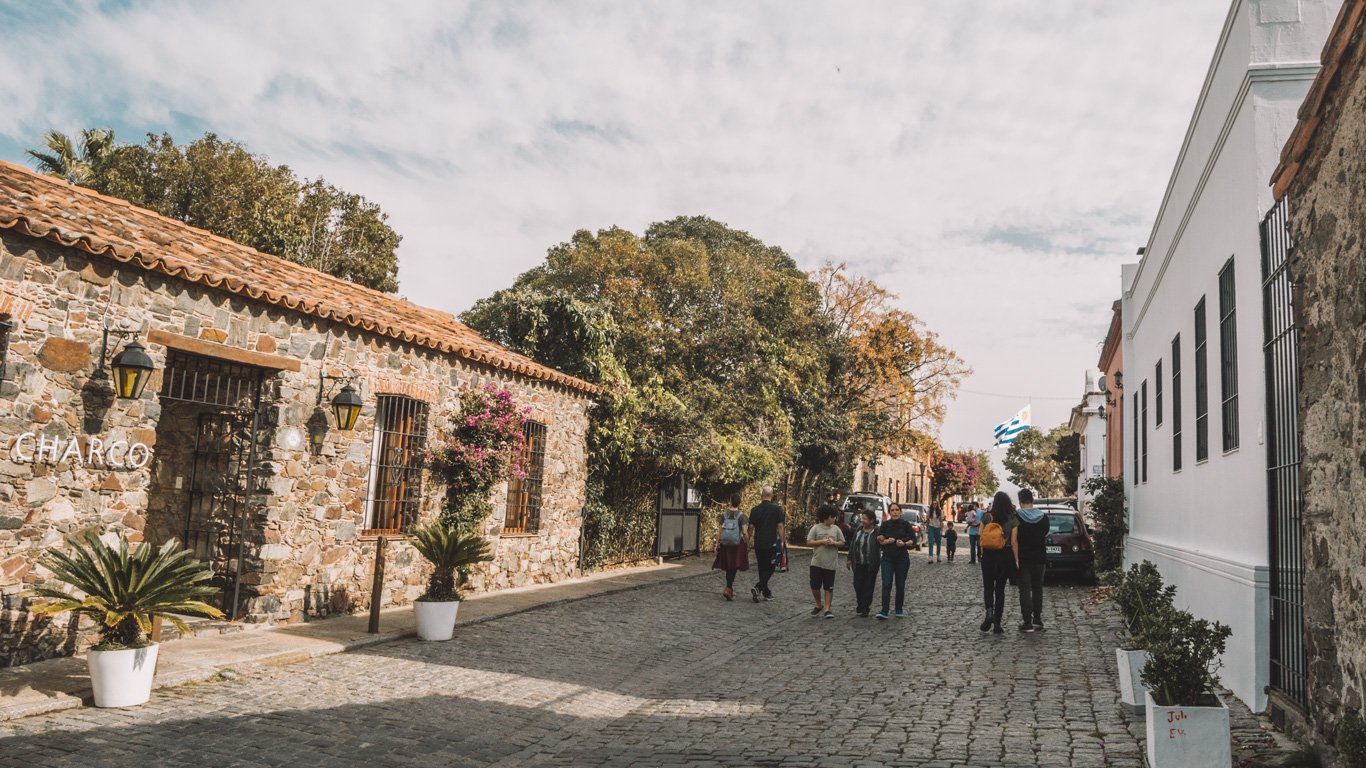 Imagem da entrada do Charco Hotel em Colonia del Sacramento, situado em uma rua de paralelepípedos movimentada.