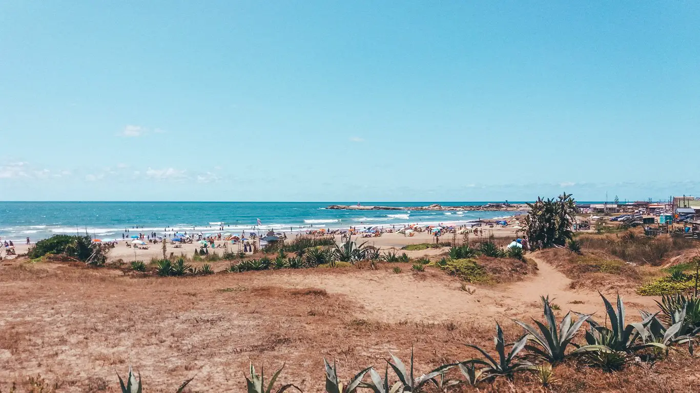 Praia de Punta del Diablo com muitas pessoas relaxando sob guarda-sóis perto da beira do mar, sob um céu azul claro e com ondas calmas. Em primeiro plano, há um terreno seco e amarronzado com plantas típicas de clima árido, como agaves, que leva até a faixa de areia movimentada e uma pequena área urbana ao fundo.