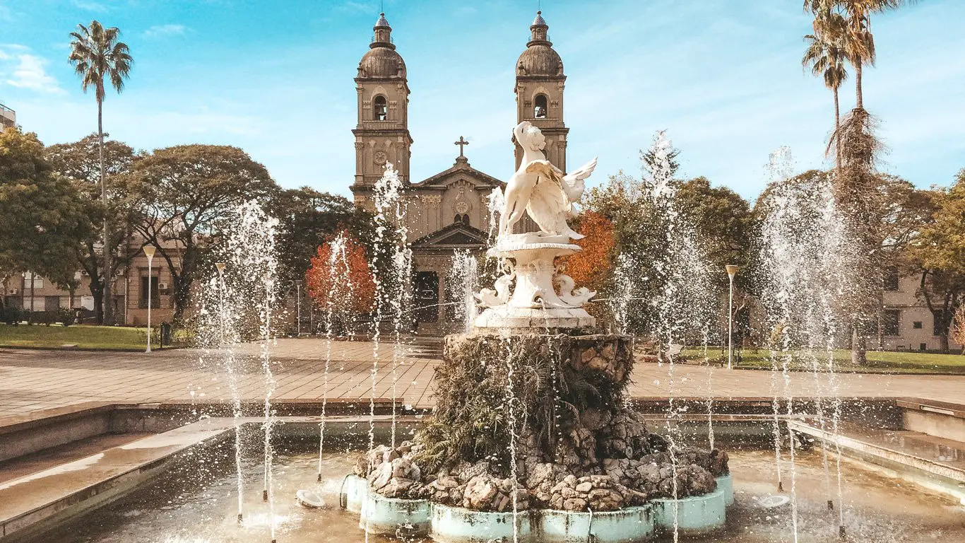 Fonte ornamentada em uma praça central de Salto, Uruguai, com esculturas clássicas cercadas por jatos d’água, emoldurada por árvores, palmeiras e uma igreja com duas torres ao fundo. A cena transmite tranquilidade e charme histórico em um ambiente urbano arborizado.