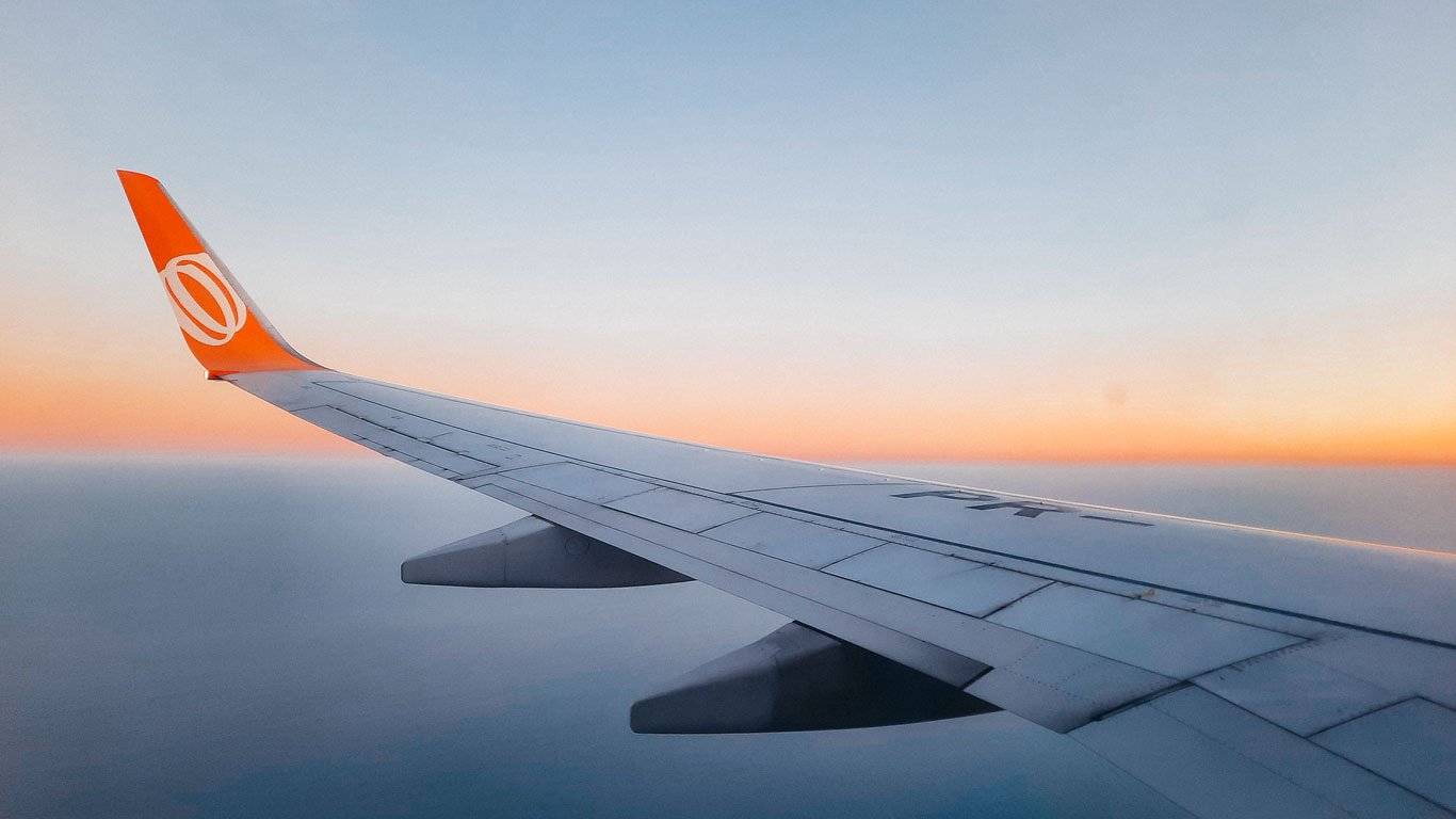 A foto mostra a asa de um avião com o logo da companhia aérea em laranja no topo, vista de dentro da cabine durante o voo. O horizonte exibe um degradê de cores suaves do amanhecer ou entardecer, variando entre azul e laranja, acima de um céu limpo.