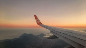 Imagem da asa de um avião com o logotipo da companhia aérea em laranja na ponta, vista durante o voo ao amanhecer ou entardecer. Abaixo, é possível ver uma paisagem costeira com montanhas e ilhas, sob um céu com tons de laranja e amarelo próximo ao horizonte.