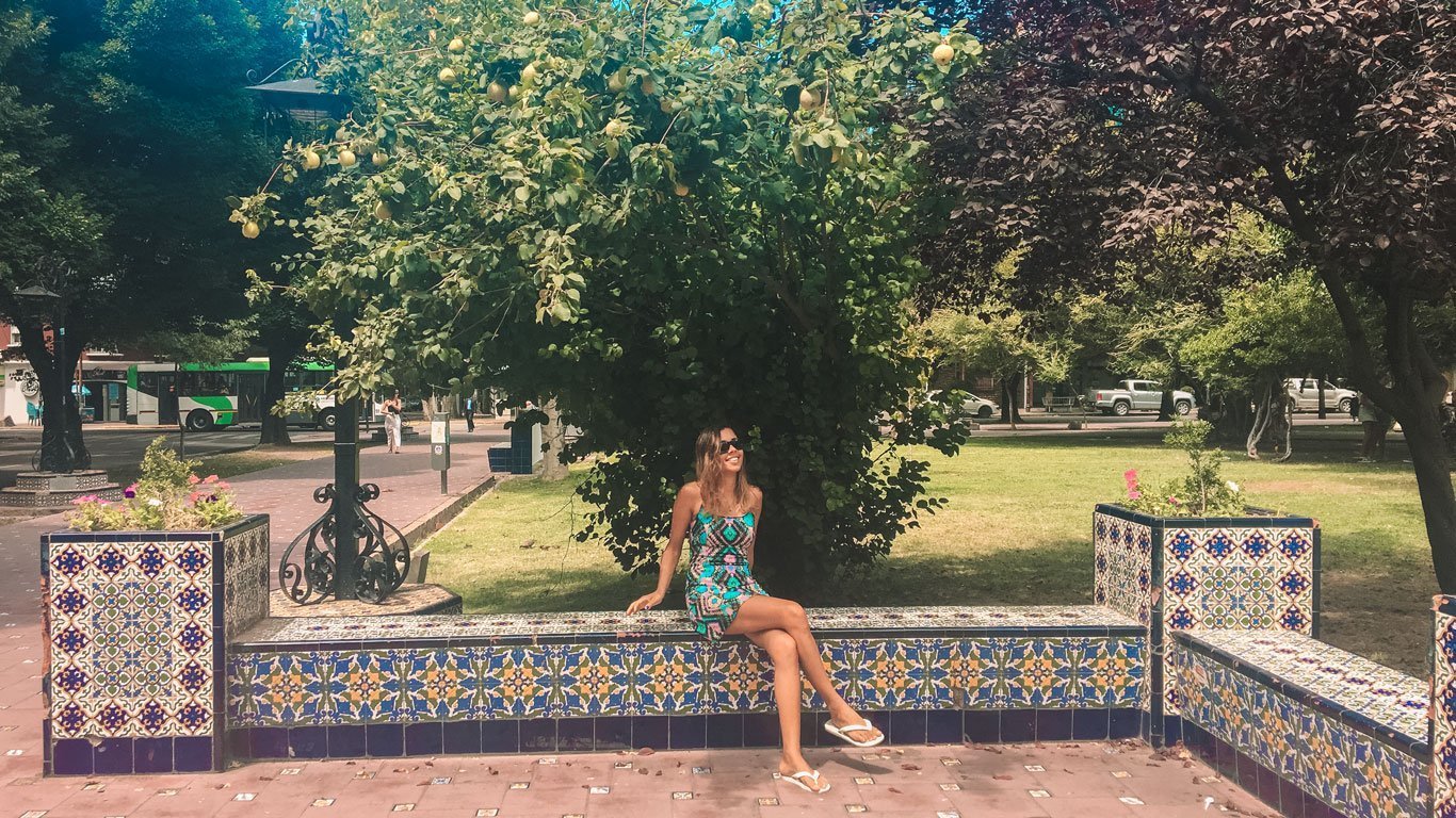 A imagem mostra uma mulher sentada em um banco de azulejos coloridos em um parque ao ar livre, com árvores ao fundo. Ela está usando um vestido estampado e óculos de sol, com um sorriso relaxado. Ao seu redor, há um ambiente arborizado, e o banco é decorado com azulejos em padrões geométricos coloridos. Ao fundo, há um ônibus verde e alguns pedestres caminhando pela praça.