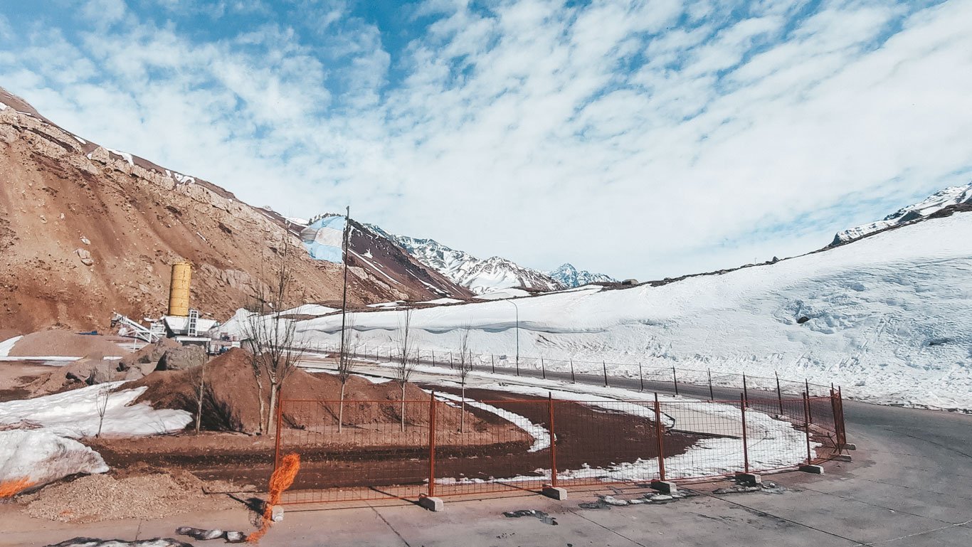 A imagem mostra uma estrada de montanha parcialmente cercada por neve, com colinas rochosas e montanhas nevadas ao fundo. O céu azul com nuvens esparsas complementa a paisagem fria e árida, e uma estrutura industrial com um grande tanque amarelo é visível à esquerda, próxima à estrada.