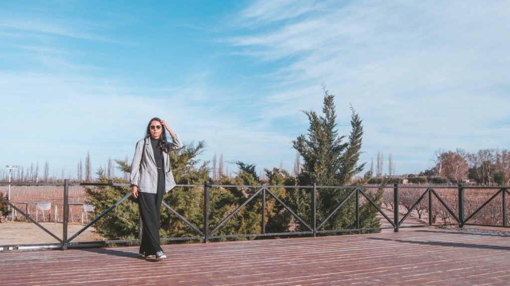 Mulher em pé sobre um deck de madeira ao ar livre, vestindo blazer xadrez cinza e calça preta, com uma mão no cabelo e usando óculos escuros. Ao fundo, uma paisagem de vinhedos e árvores contra um céu azul claro, com cercas e árvores esparsas que adicionam profundidade à cena natural.