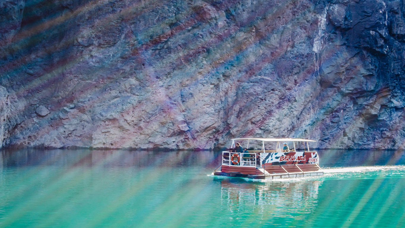 A imagem mostra um barco turístico navegando em águas cristalinas de um tom verde-azulado, próximo a uma parede rochosa imponente. No barco, há algumas pessoas sentadas sob uma cobertura. A luz do sol cria um efeito de arco-íris nas rochas e na água, adicionando um toque colorido à paisagem natural.