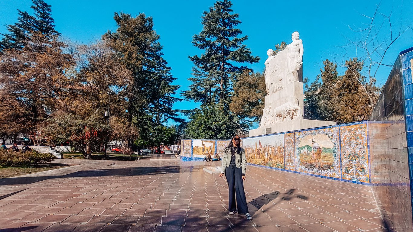 Mulher posando para uma foto na Plaza España, Centro de Mendoza. Ao fundo um grande monumento e árvores.