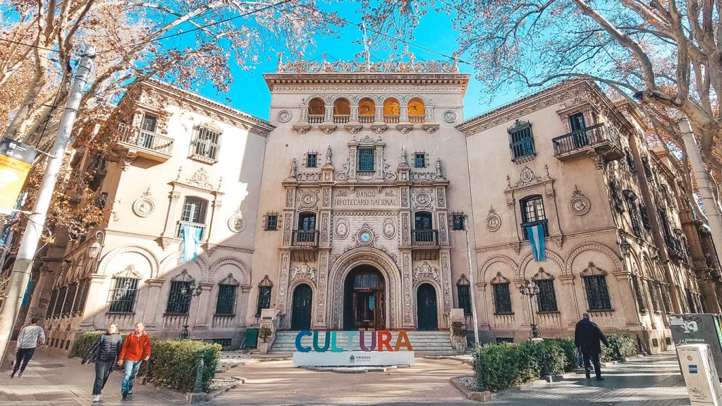 Imagem mostrando um edifício histórico em Mendoza, Argentina, com uma fachada detalhada, onde se lê “Banco Hipotecario Nacional” no topo. Na frente, há a palavra “CULTURA” em letras coloridas, e uma bandeira azul e branca está pendurada na entrada. Pessoas caminham em um dia claro, com árvores sem folhas ao redor, indicando possivelmente uma estação mais fria.