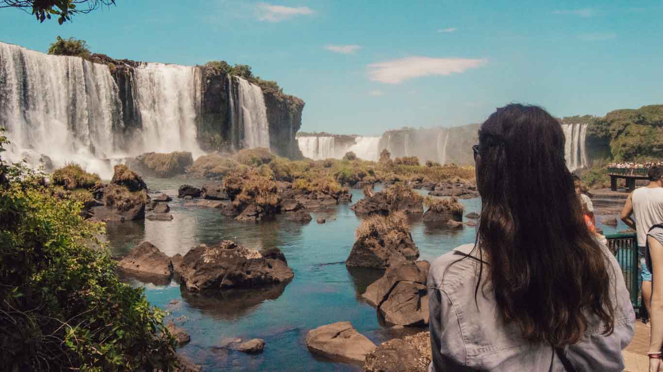 Mulher de cabelos longos e de costas, admirando as quedas de água das Cataratas do Iguaçu, no Brasil.