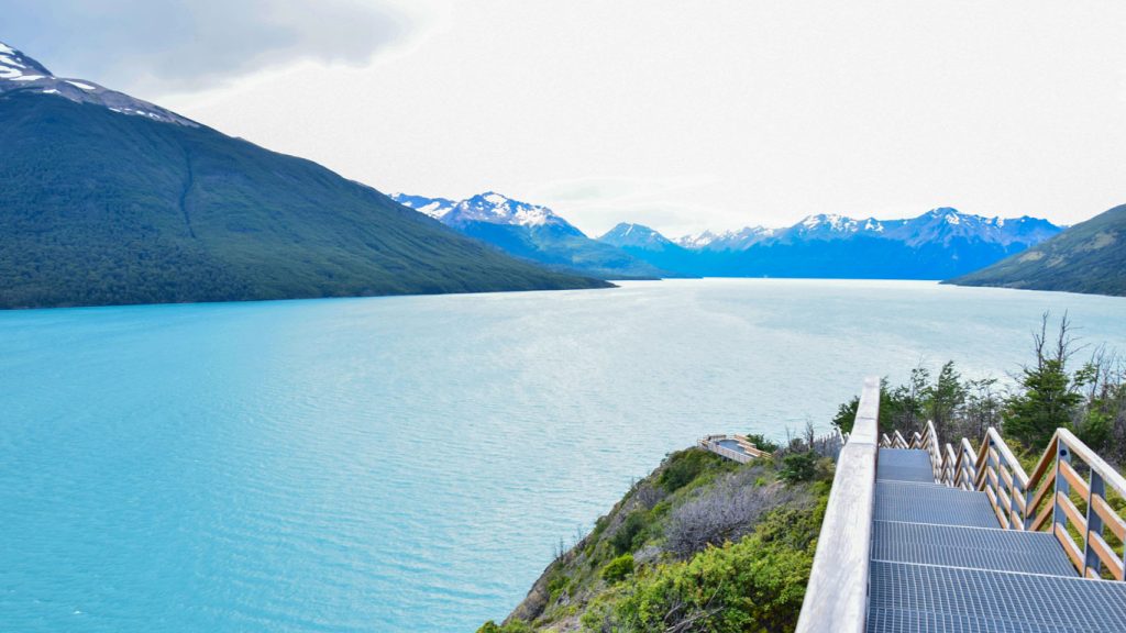 Uma passarela em madeira, margeando um lago com águas azuis e envolto por montanhas nevadas, em El Calafate, na Argentina.