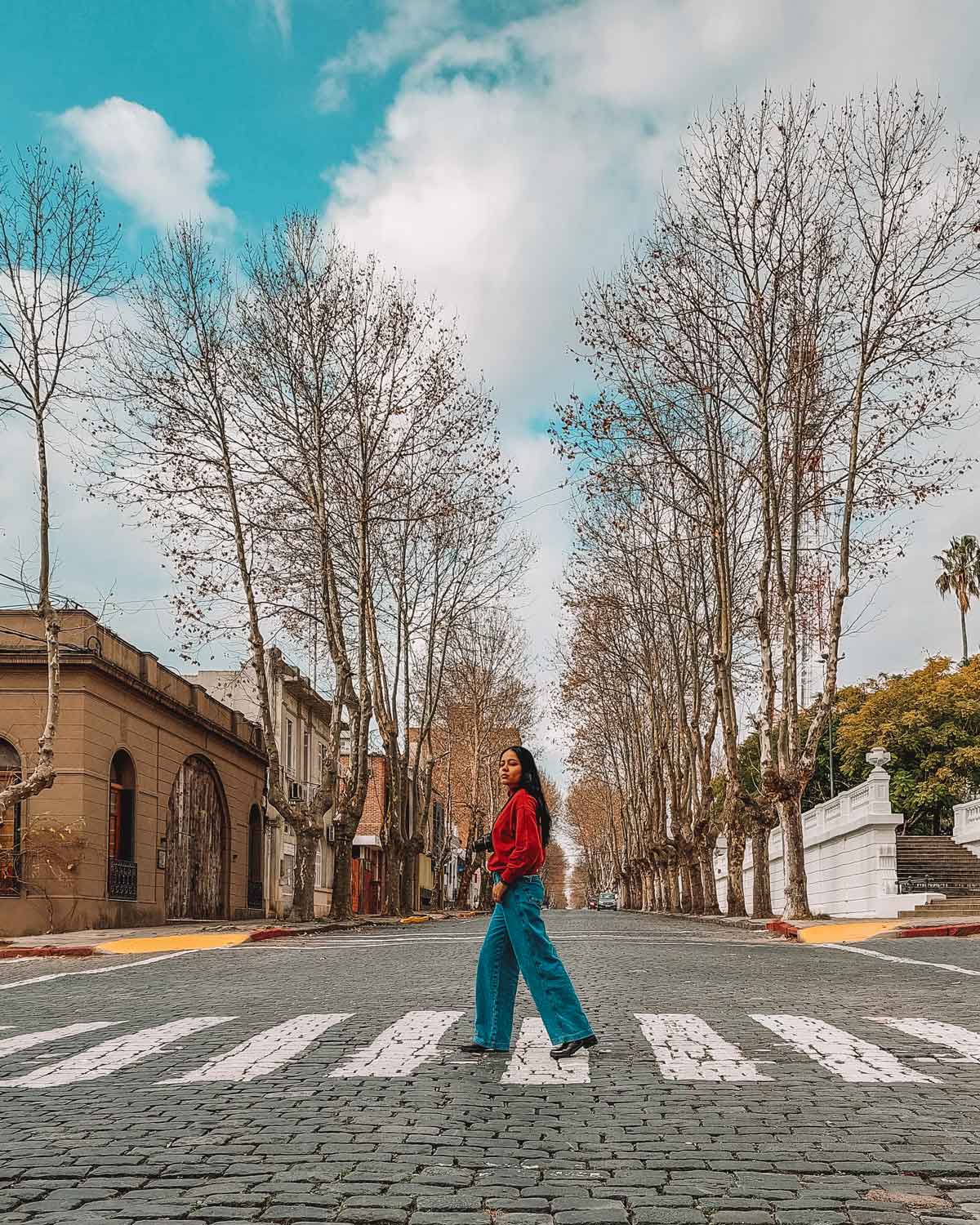 Mulher com uma blusa vermelhas e calça jeans atravessando uma rua de pedras em Colonia del Sacramento, Uruguai.