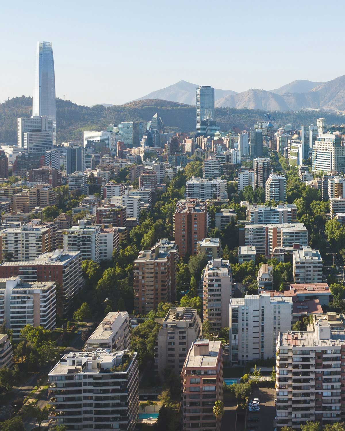 bairro-providencia-santiago Vista aérea de Providência, uma área moderna e arborizada de Santiago do Chile, com diversos prédios residenciais e comerciais. Ao fundo, destaca-se o icônico edifício Costanera Center com as montanhas dos Andes ao longe.