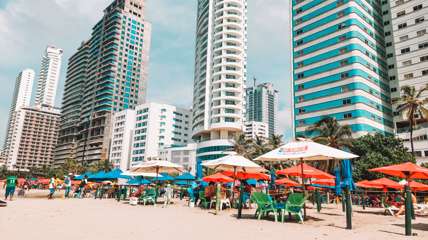 Praia de Bocagrande, em Cartagena. A praia tem areia amarelada e está cheia de Guarda Sol, ao fundo os prédios modernos do bairro.