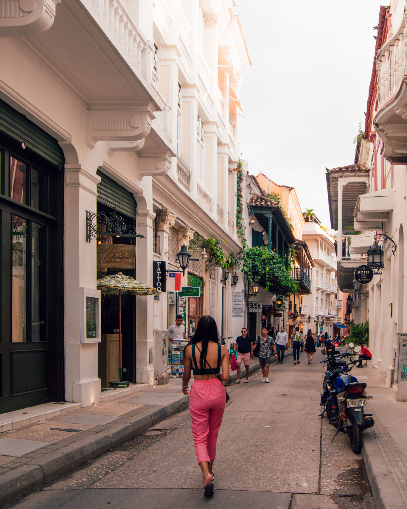 Mulher caminhando pelas ruas estreitas do centro de Cartagena, na Colômbia. A mulher está de costas e veste unha calça rosa e uma blusa preta, seus cabelos são longos e pretos e ela caminha despretensiosamente. As ruas são acompanhadas de casas brancas, com portas coloridas e plantas verdinhas nas varandas.