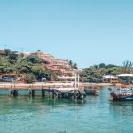 Imagem de uma paisagem costeira de Búzios, Brasil, com barcos de pesca ancorados em um píer de madeira sobre águas cristalinas. Ao fundo, uma colina coberta por vegetação e construções residenciais de diferentes tamanhos e cores, com um céu azul claro acima. A atmosfera é tranquila, destacando a beleza natural e o charme rústico da cidade.