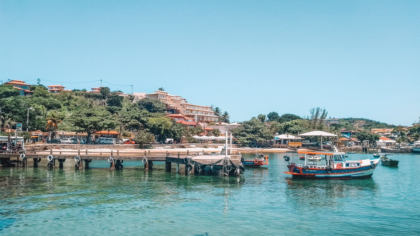 Imagem de uma paisagem costeira de Búzios, Brasil, com barcos de pesca ancorados em um píer de madeira sobre águas cristalinas. Ao fundo, uma colina coberta por vegetação e construções residenciais de diferentes tamanhos e cores, com um céu azul claro acima. A atmosfera é tranquila, destacando a beleza natural e o charme rústico da cidade.