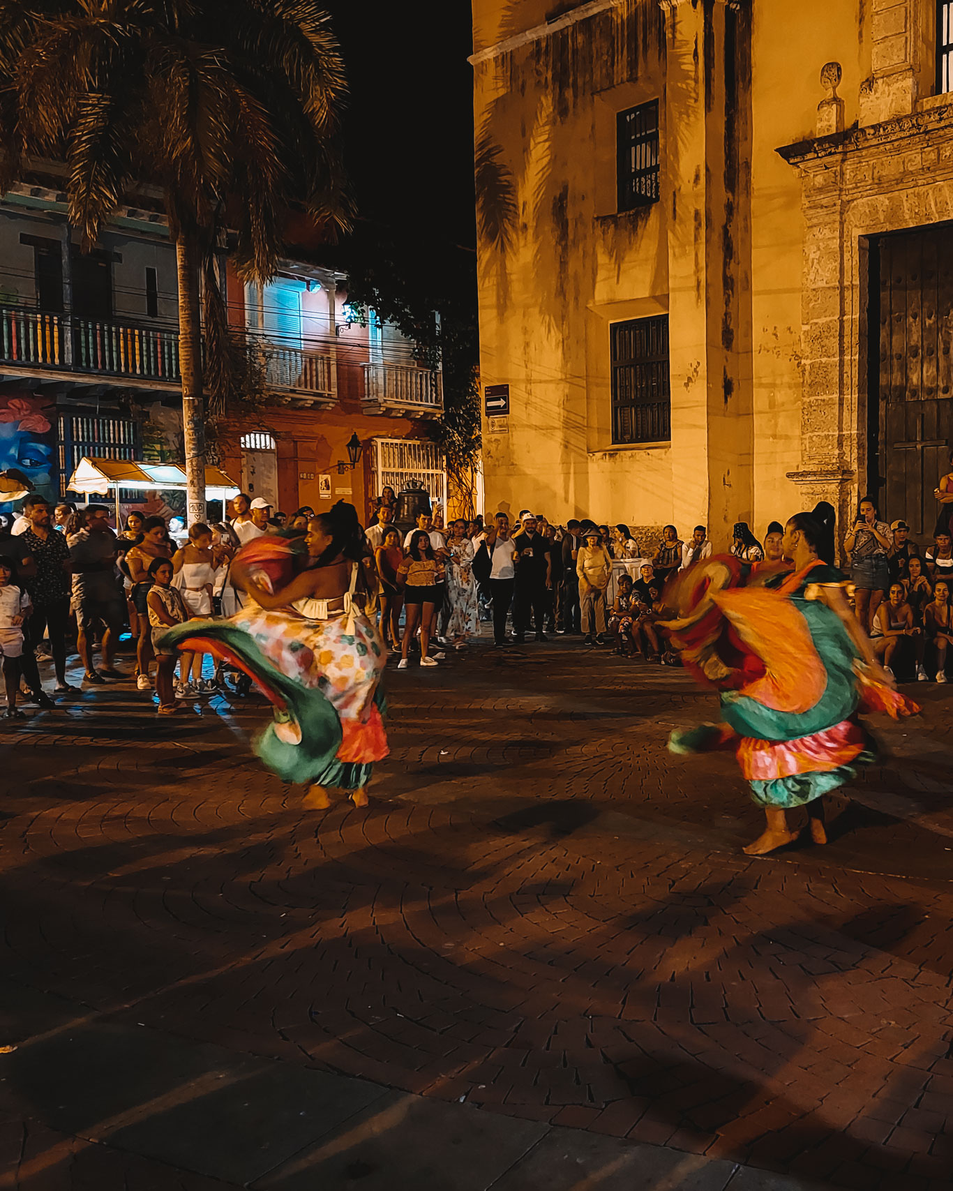 O bairro Getsemaní durante a noite, com bailarinas vestindo roupas caribenhas rodadas e coloridas, dançando pela praça do bairro.