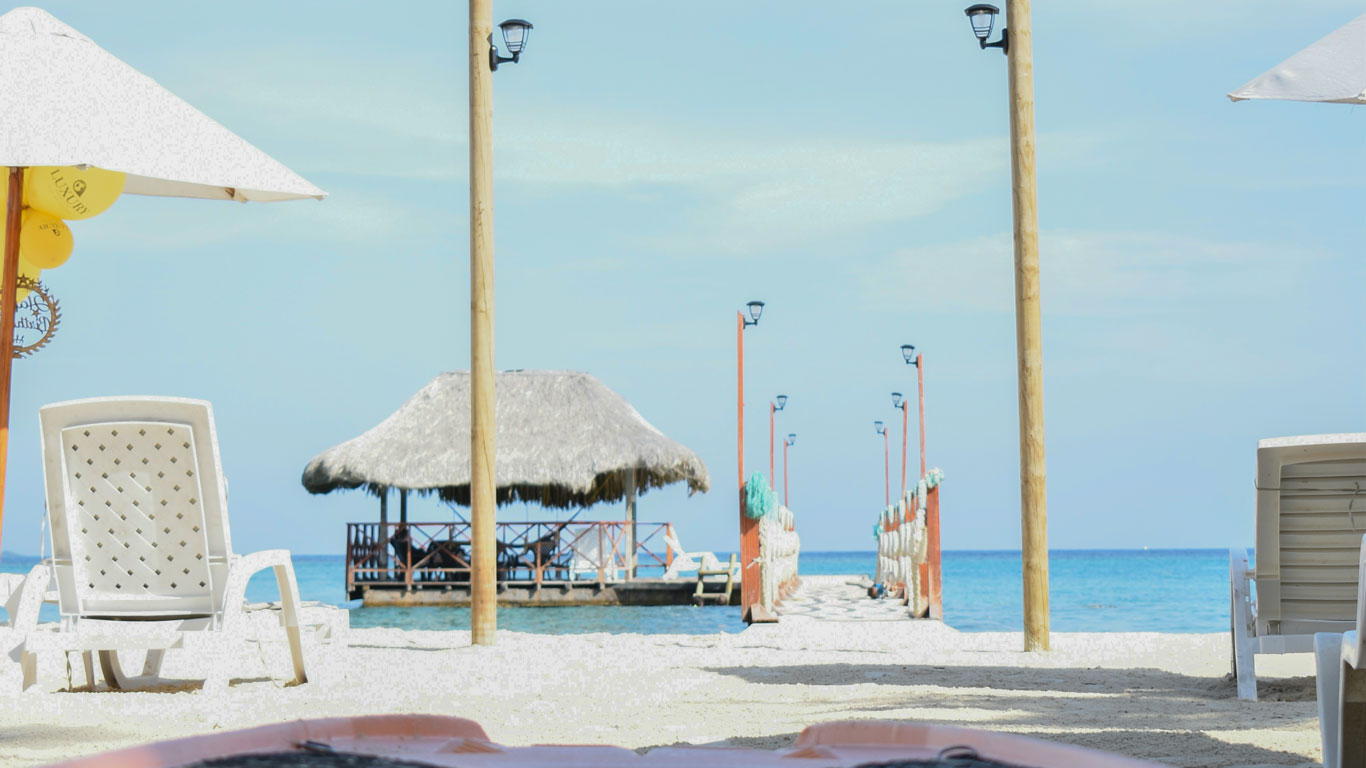 Entrada do resort Luxury Beach Club em Islas del Rosario, Cartagena. Em primeiro plano um píer de madeira com uma tenda coberta ao fianal, ao fundo o mar azul se funde com o céu.