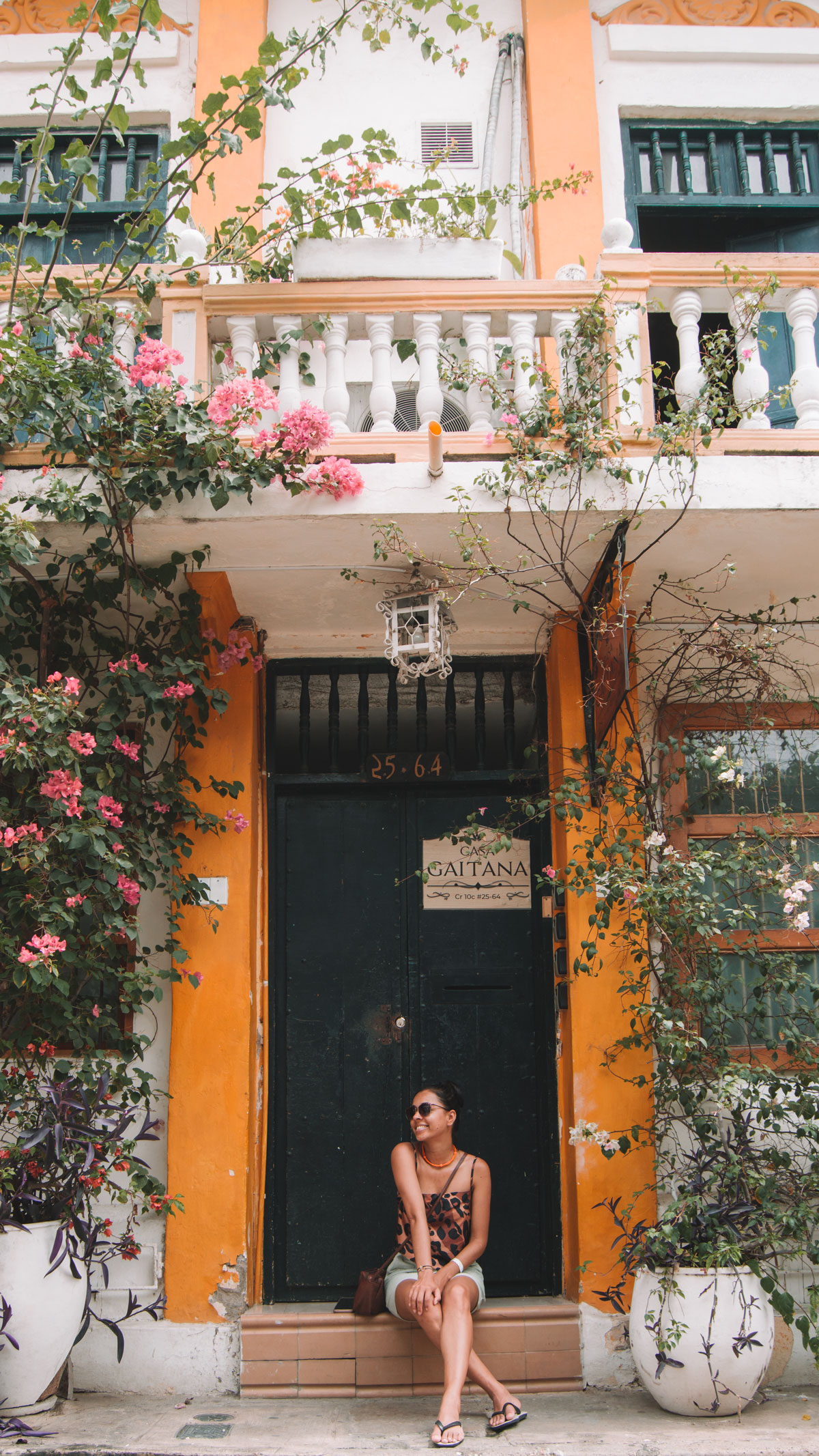 Mulher sentada com as pernas cruzadas e mão apoiadas sobre as penas, em uma parta amarela e verde do bairro Getsemaní em Cartagena. A mulher está de óculos escuro e cabelo preso.