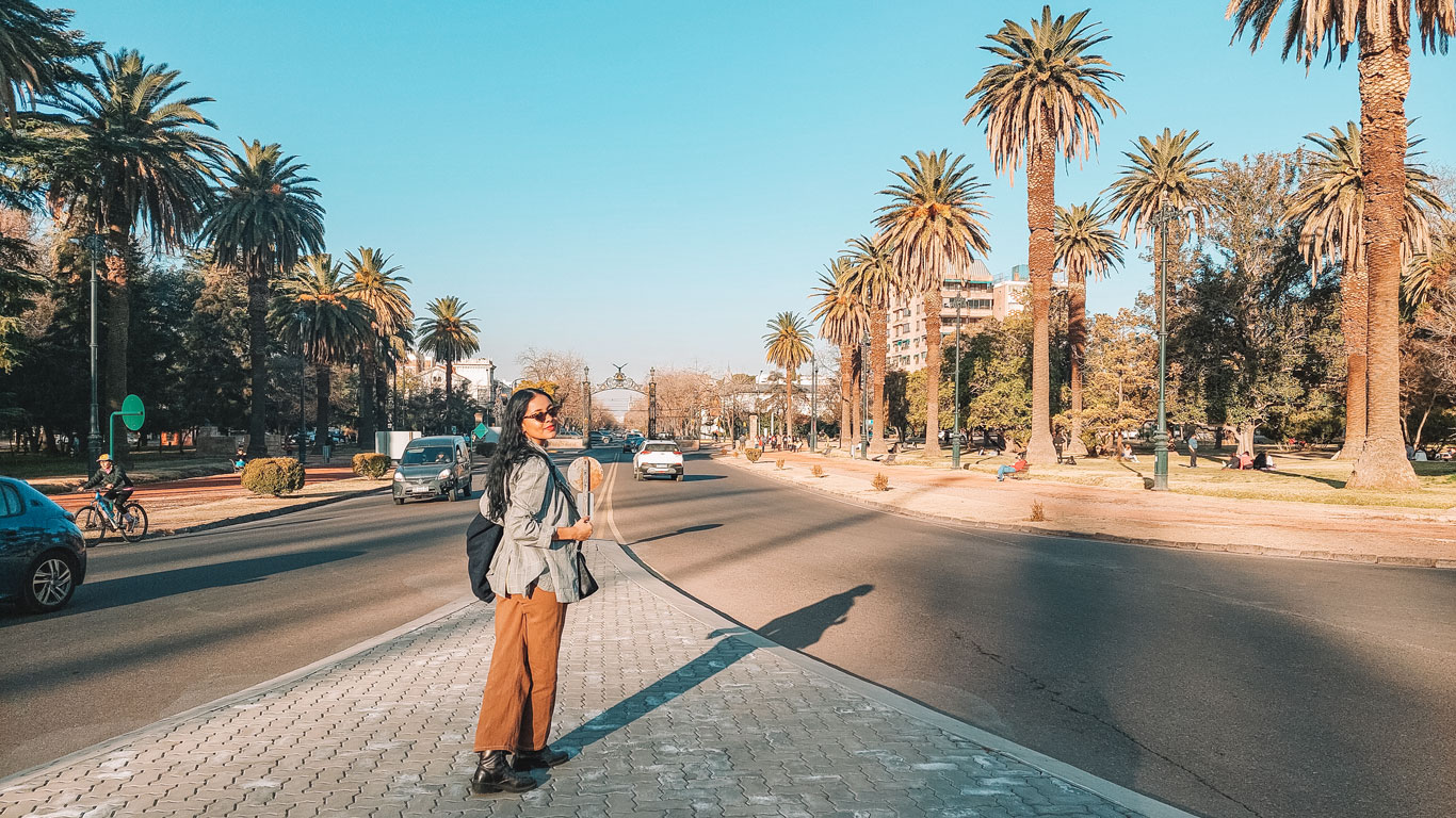 Mulher caminhando em frente a entrada do Parque General San Martin em Mendoza. A mulher de cabelos longos e escuros, está de calça jeans marrom, blazer cinza e óculos escuro.