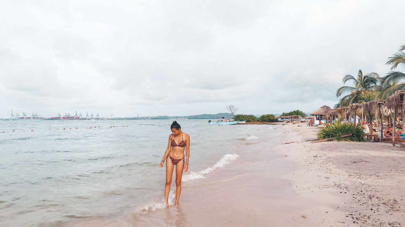 Mulher usando um biquíni roxo e caminhando na praia do Hotel Fenix Beach na Ilha Tierra Bomba. Ao fundo, pequenas tendas com camas de praia, acompanham a orla.